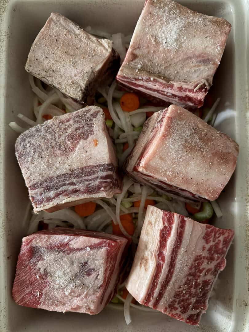 Six raw, seasoned beef short ribs are arranged in a baking dish on top of sliced onions, carrots, and celery, ready for roasting or braising.