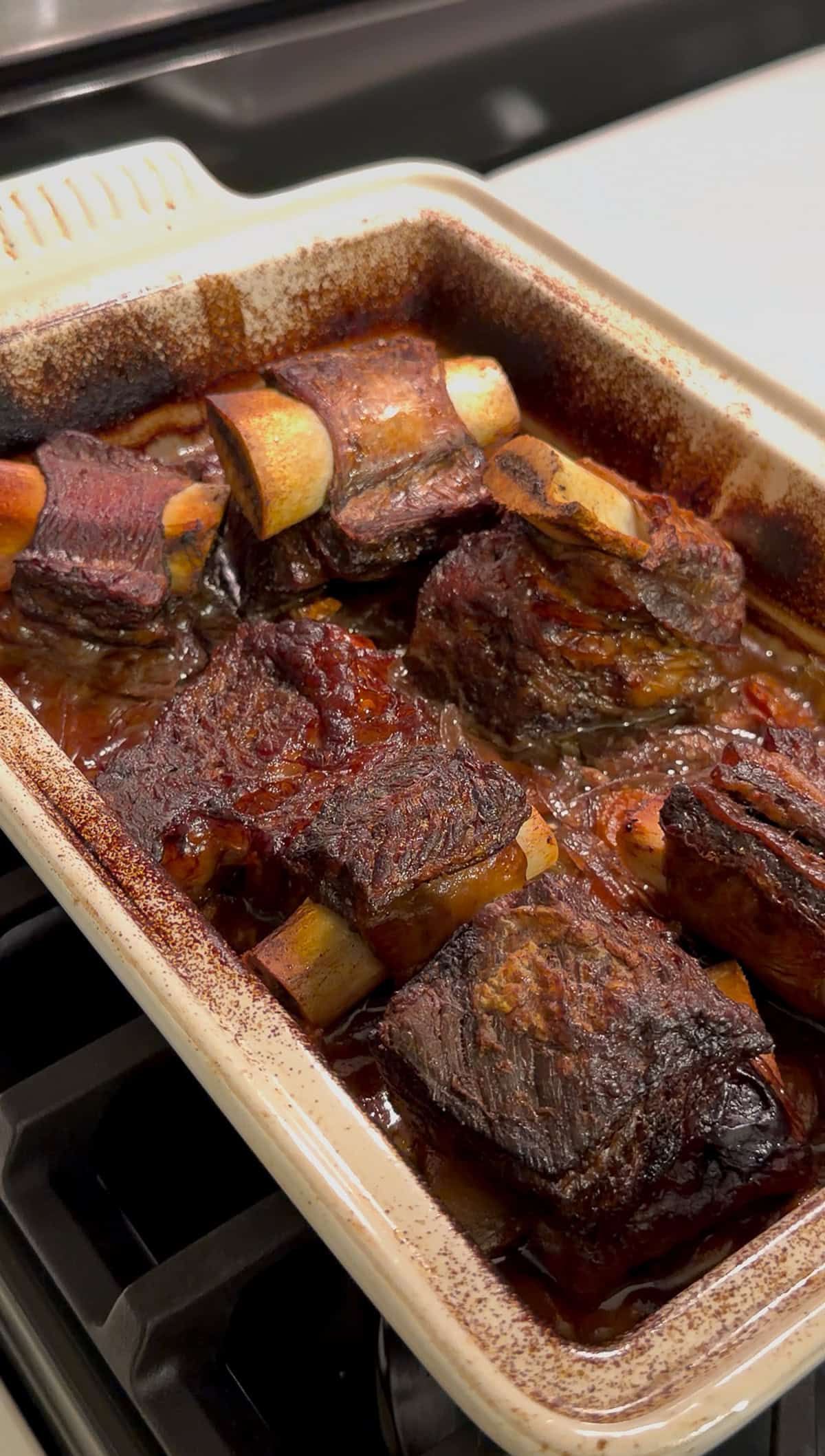 Braised beef short ribs in a rectangular baking dish, cooked until dark brown with a caramelized crust, sitting in a rich, savory sauce on a stovetop.