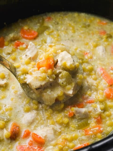 A close-up of a ladle lifting a serving of thick split pea soup with chunks of carrots, celery, and pieces of meat from a pot. The soup appears hearty and creamy with visible vegetables.