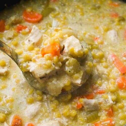 A close-up of a ladle lifting a serving of thick split pea soup with chunks of carrots, celery, and pieces of meat from a pot. The soup appears hearty and creamy with visible vegetables.