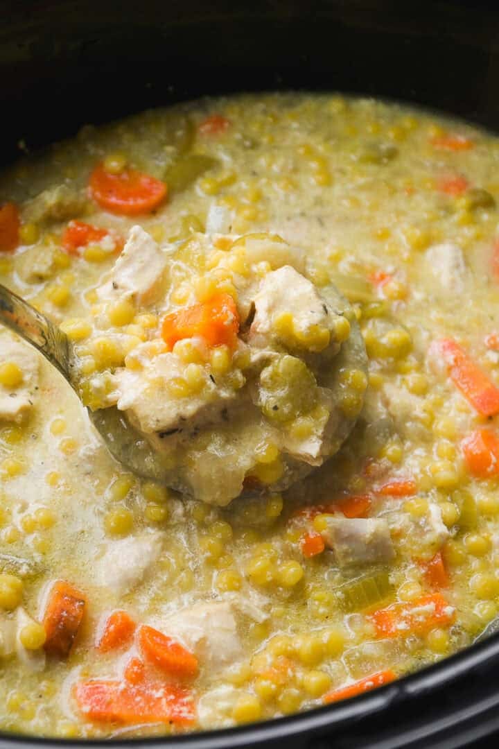 A close-up of a ladle lifting a serving of thick split pea soup with chunks of carrots, celery, and pieces of meat from a pot. The soup appears hearty and creamy with visible vegetables.