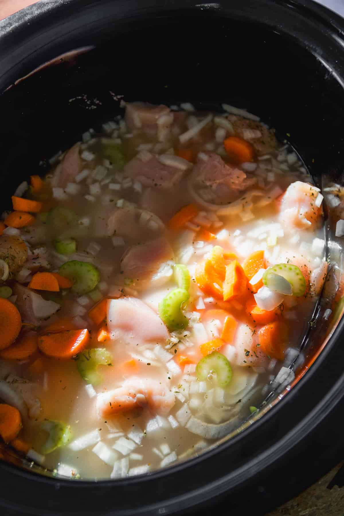 Raw chicken, chopped carrots, celery, and onions in broth inside a black slow cooker, ready to be cooked. Sunlight illuminates part of the ingredients.