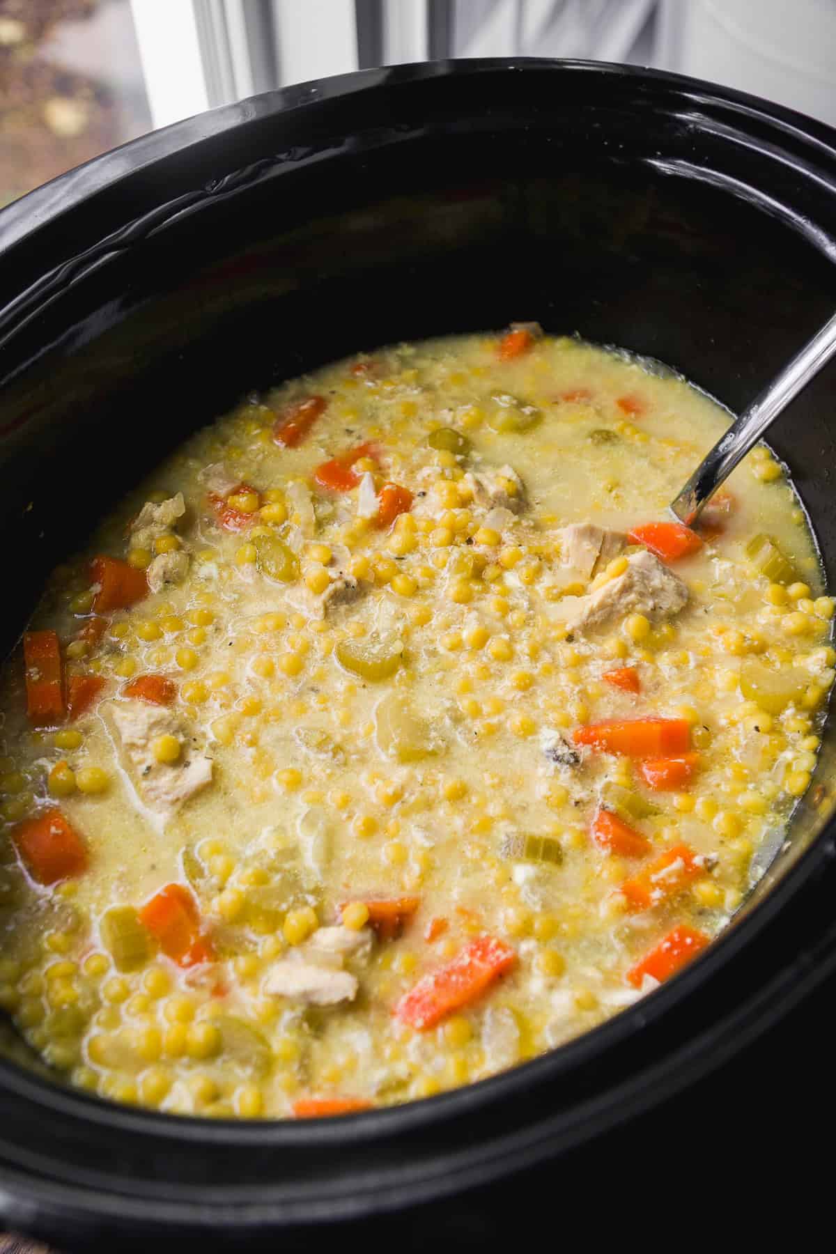 A close-up of a crockpot filled with chicken soup containing carrots, celery, tiny round pasta, and chunks of chicken in a creamy broth, with a metal spoon inside.
