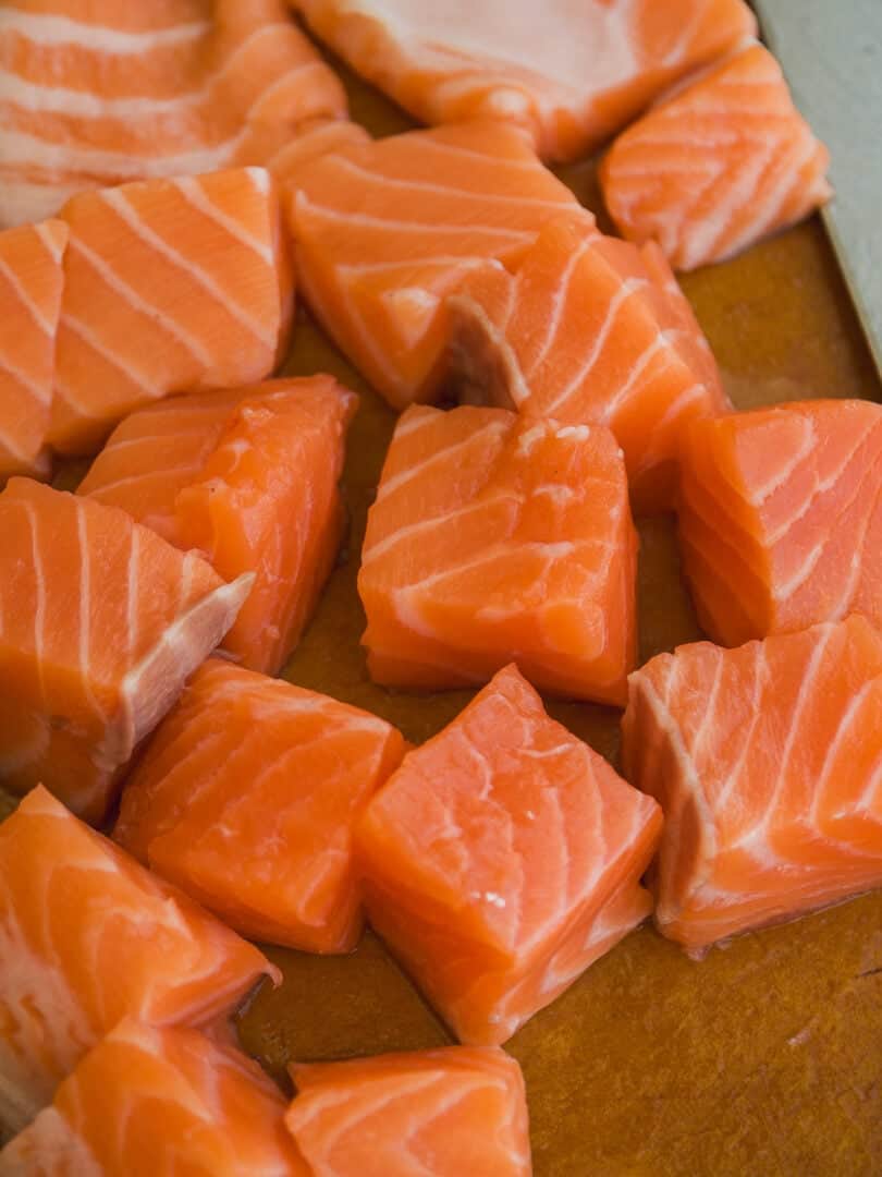 Chunks of raw salmon with visible white marbling are arranged on a wooden cutting board, next to the edge of a metal knife.
