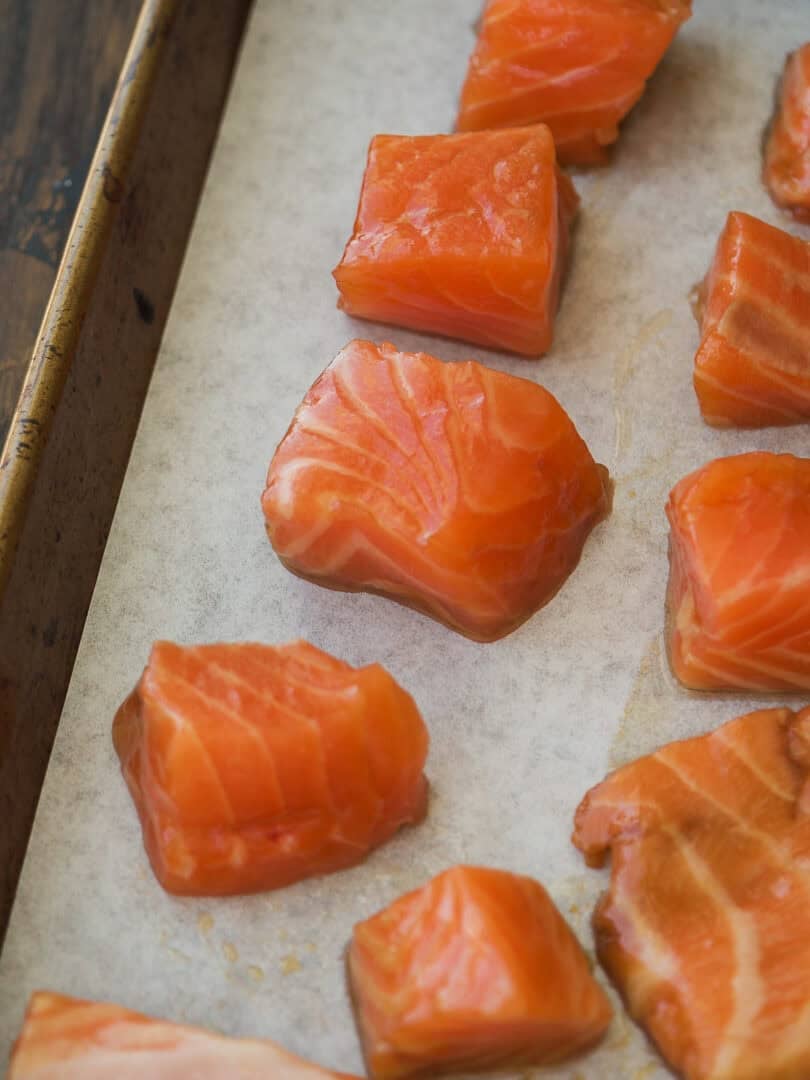 Raw salmon chunks are arranged on a parchment-lined baking sheet, ready for cooking. The fish pieces display a bright orange color and marbled texture.
