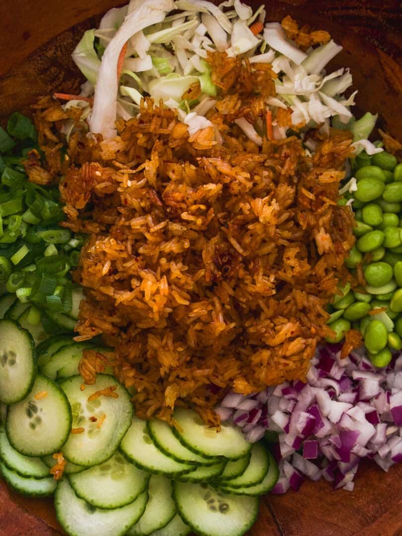A bowl filled with sections of cooked rice, sliced cucumber, shredded cabbage, chopped green onions, diced red onion, and shelled edamame arranged in a colorful, appetizing display.