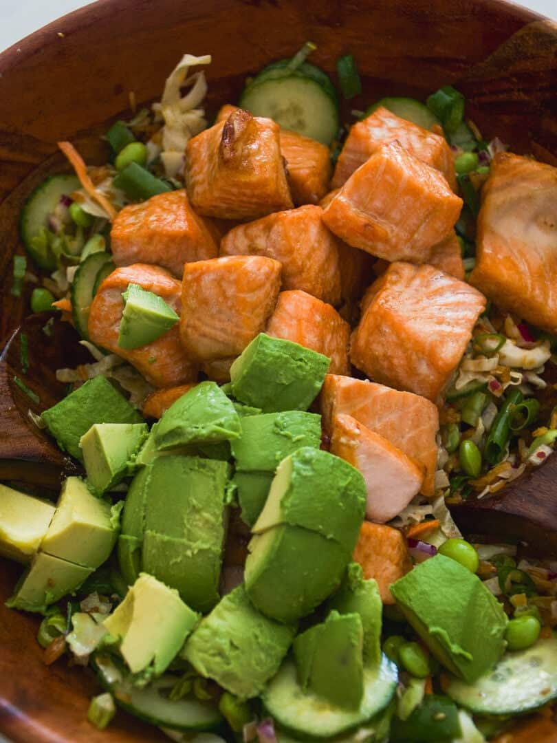 A wooden bowl filled with chunks of cooked salmon, sliced avocado, cucumber, shredded cabbage, and green onions, creating a colorful and fresh salad.
