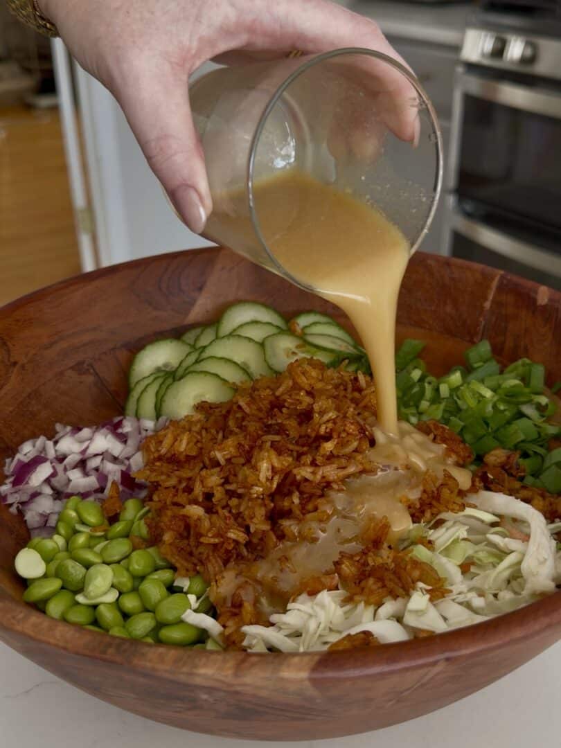 A hand pours creamy dressing from a glass onto a wooden bowl filled with chopped vegetables, including cucumber, red onion, edamame, green onion, cabbage, and crispy fried onions.