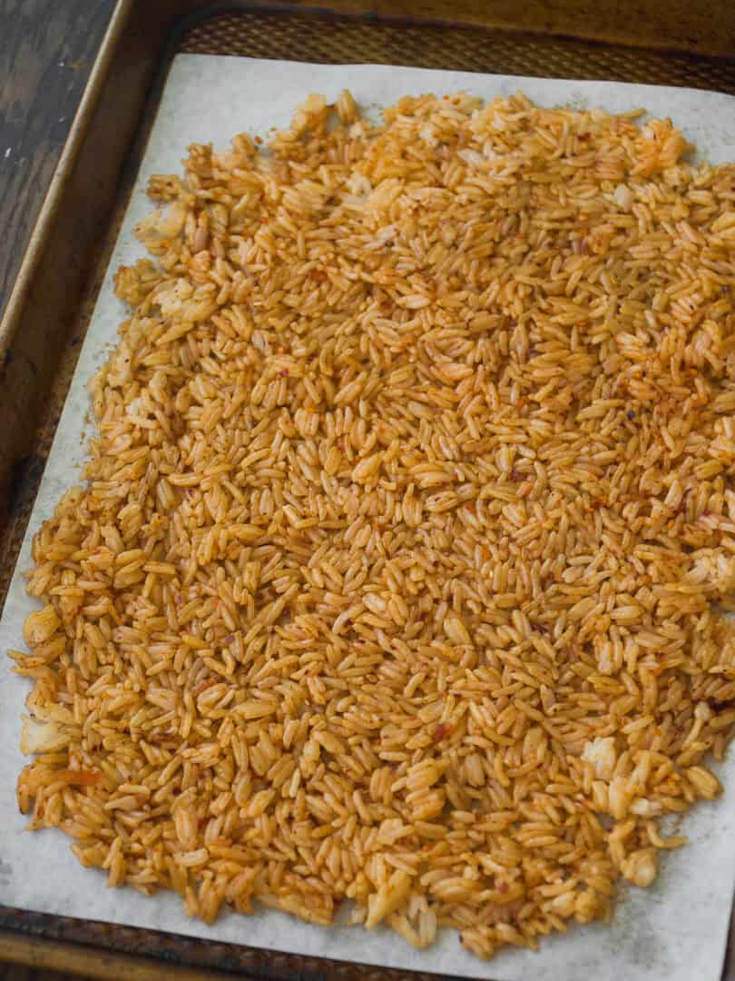 A baking tray lined with parchment paper, spread with a single layer of cooked, reddish-orange rice, likely seasoned or baked, on a wooden surface.