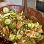A hand holds a wooden bowl filled with a colorful salad containing sliced cucumbers, avocado, edamame, cabbage, red onion, and pieces of seasoned salmon, with a wooden spoon ready for serving.