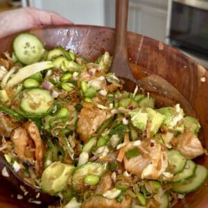 A hand holds a wooden bowl filled with a colorful salad containing sliced cucumbers, avocado, edamame, cabbage, red onion, and pieces of seasoned salmon, with a wooden spoon ready for serving.