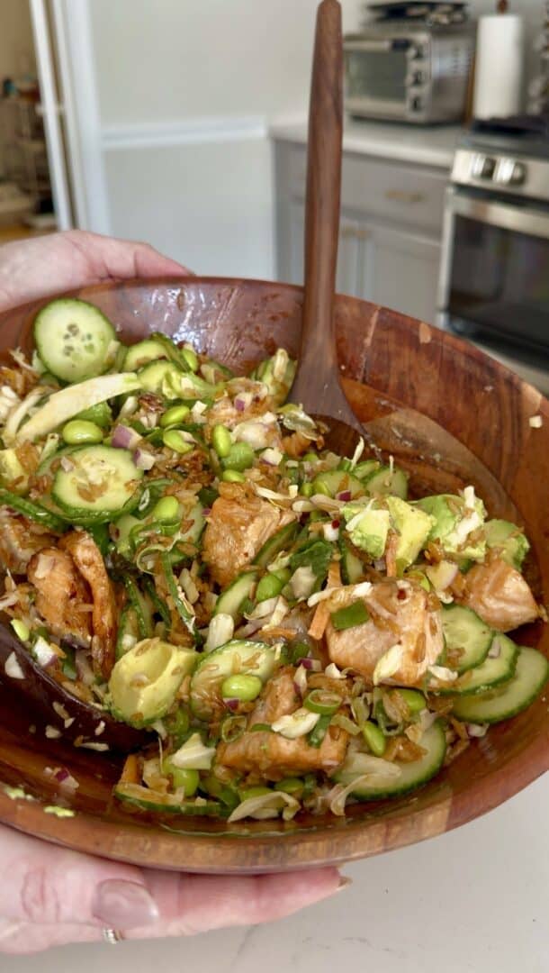 A hand holds a wooden bowl filled with a colorful salad containing sliced cucumbers, avocado, edamame, cabbage, red onion, and pieces of seasoned salmon, with a wooden spoon ready for serving.