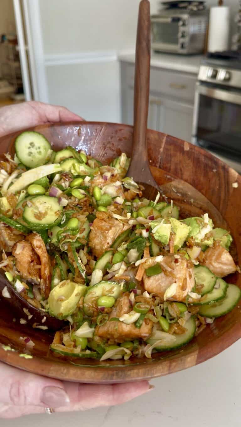A hand holds a wooden bowl filled with a colorful salad containing sliced cucumbers, avocado, edamame, cabbage, red onion, and pieces of seasoned salmon, with a wooden spoon ready for serving.