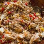 A wooden bowl filled with a hearty grain salad featuring chunks of tofu, diced red and yellow bell peppers, and red onion—similar in style to a halloumi cheese salad—all mixed together with a spoon.
