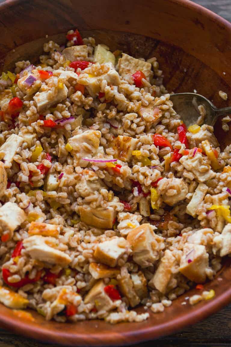 A wooden bowl filled with a hearty grain salad featuring chunks of tofu, diced red and yellow bell peppers, and red onion—similar in style to a halloumi cheese salad—all mixed together with a spoon.