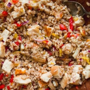 A close-up of a bowl filled with a mixed grain salad containing diced chicken, red bell peppers, yellow bell peppers, red onions, and cooked brown rice or barley, with a spoon partially visible.