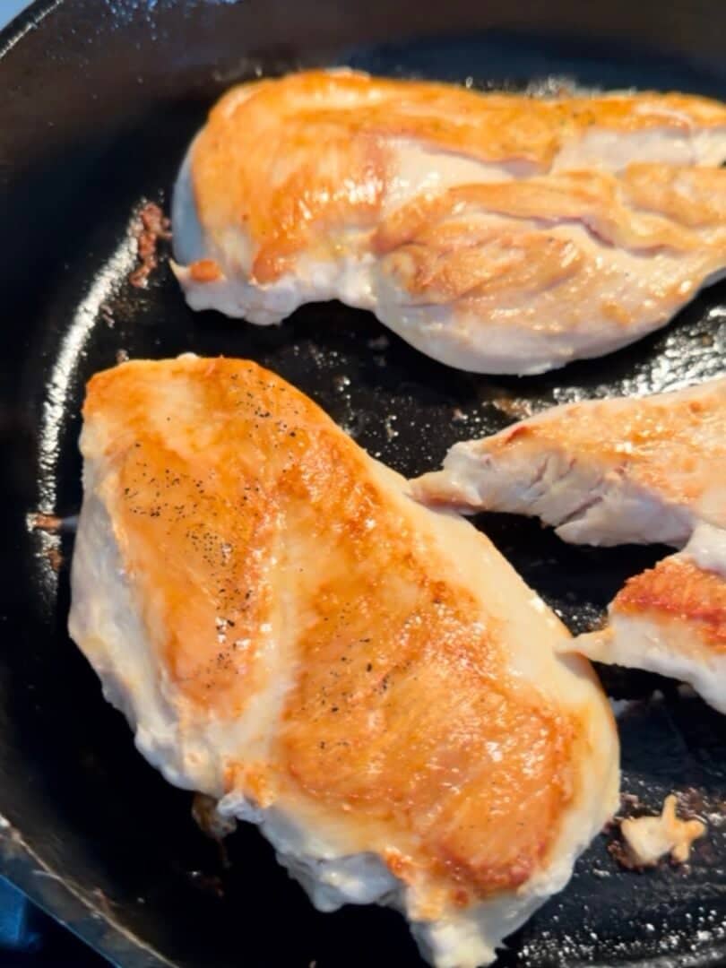Three pieces of chicken breast are searing in a black cast iron skillet, showing a golden brown crust on the surface. The skillet is on a stove, and the chicken appears to be cooking evenly.