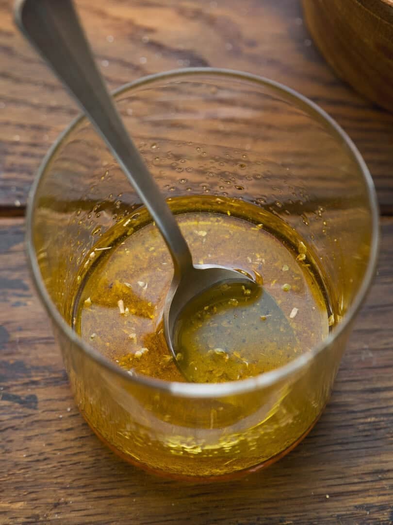 A glass with a metal spoon contains a golden liquid, likely olive oil and herbs, sitting on a wooden surface.