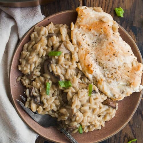 A plate with creamy mushroom orzo garnished with green onions, next to a piece of seared chicken breast. A fork rests on the plate, and part of a cooking pan and napkin are visible on a rustic wooden table.