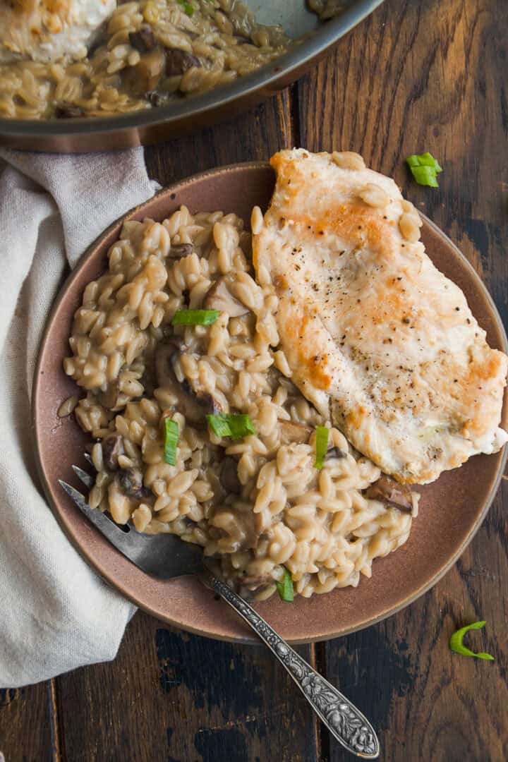 A plate with creamy mushroom orzo garnished with green onions, next to a piece of seared chicken breast. A fork rests on the plate, and part of a cooking pan and napkin are visible on a rustic wooden table.