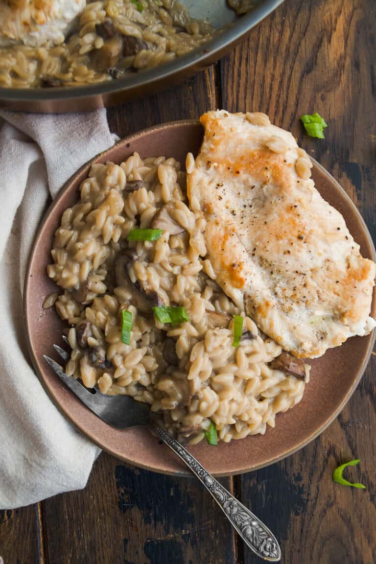 A plate with creamy mushroom orzo garnished with green onions, next to a piece of seared chicken breast. A fork rests on the plate, and part of a cooking pan and napkin are visible on a rustic wooden table.