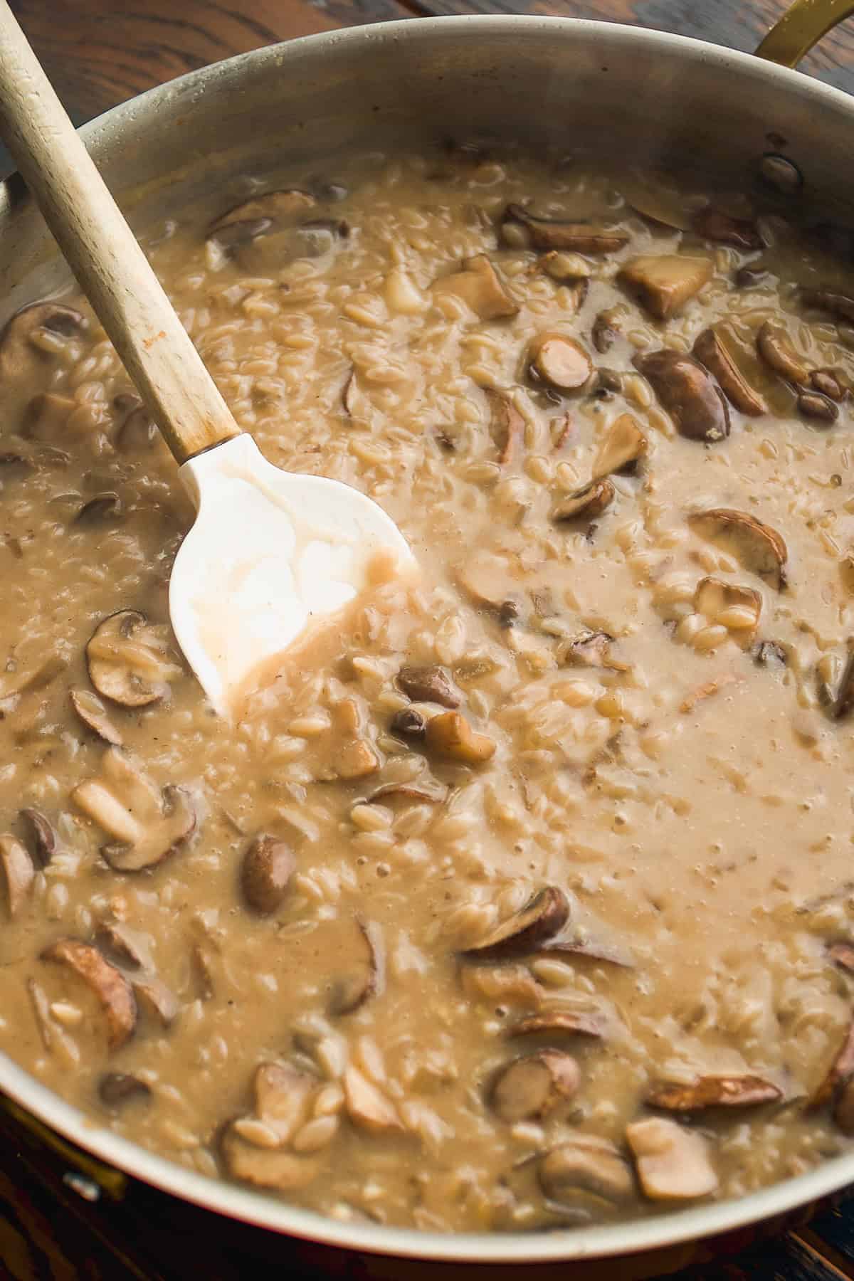A creamy mushroom risotto is being stirred in a large pan with a white spatula. Sliced mushrooms and rice are visible throughout the thick, rich mixture.