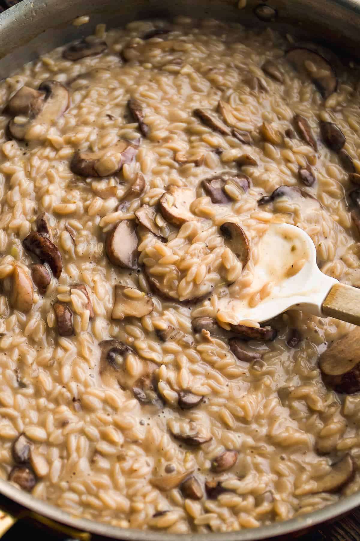 A close-up of creamy mushroom risotto being stirred with a white spatula in a skillet. The rice is tender and coated in a rich, sauce with visible slices of cooked mushrooms.