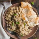 A plate with creamy mushroom orzo topped with chopped green onions and a piece of seasoned, cooked chicken breast, set on a rustic wooden table beside a white napkin. A fork rests on the plate.