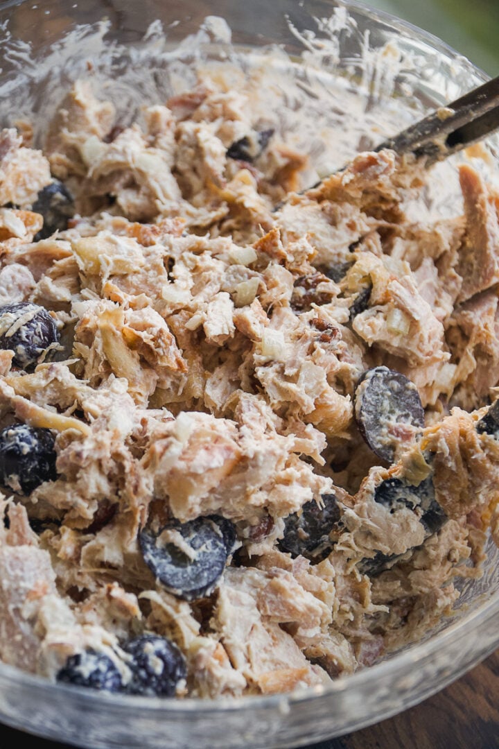 A close-up of a glass bowl filled with rotisserie chicken salad, mixed with halved black grapes and a creamy dressing, with a spoon partially visible in the mixture.
