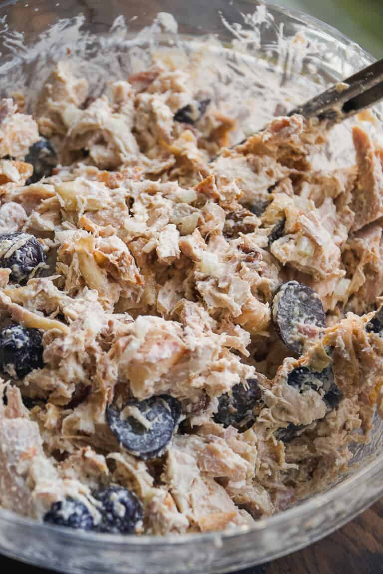 A close-up of a glass bowl filled with rotisserie chicken salad, mixed with halved black grapes and a creamy dressing, with a spoon partially visible in the mixture.