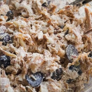 A close-up of a bowl filled with chicken salad, featuring shredded chicken, halved grapes, and a creamy dressing being mixed with utensils.