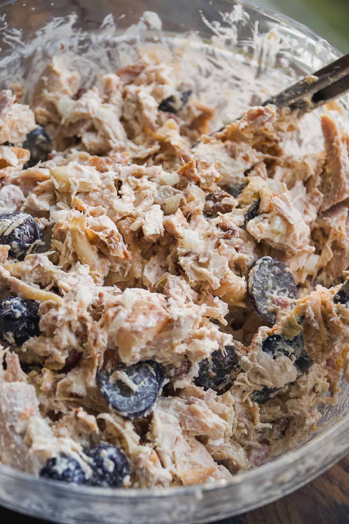 A close-up of a bowl filled with chicken salad, featuring shredded chicken, halved grapes, and a creamy dressing being mixed with utensils.