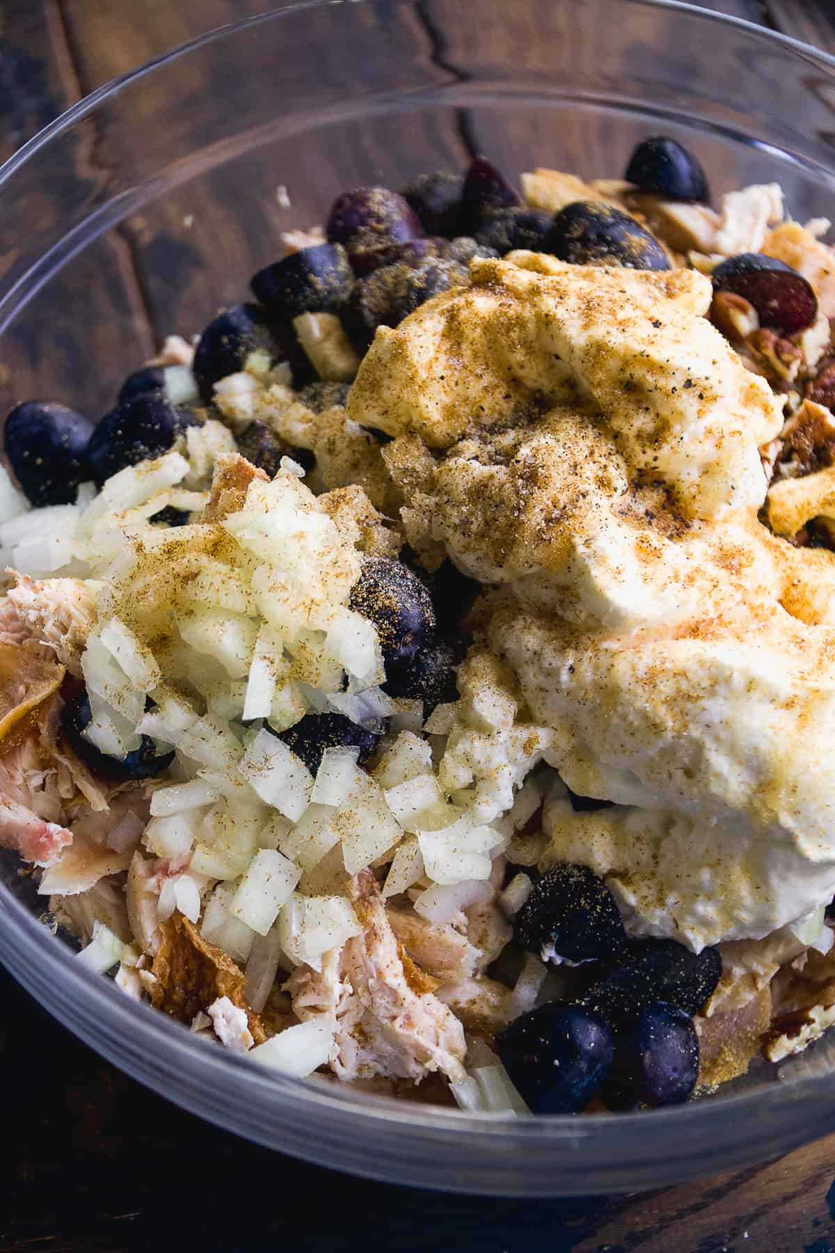 A clear bowl filled with chopped onions, shredded chicken, dark grapes, and a generous dollop of creamy mayonnaise topped with black pepper and seasoning, ready to be mixed into a salad.