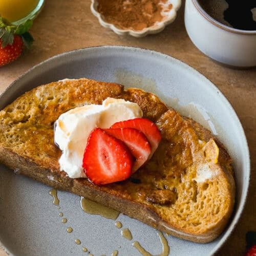 A plate with a slice of French toast topped with whipped cream and sliced strawberries, drizzled with syrup. Nearby are a cup of coffee, a glass of orange juice, strawberries, and a small dish of cinnamon.