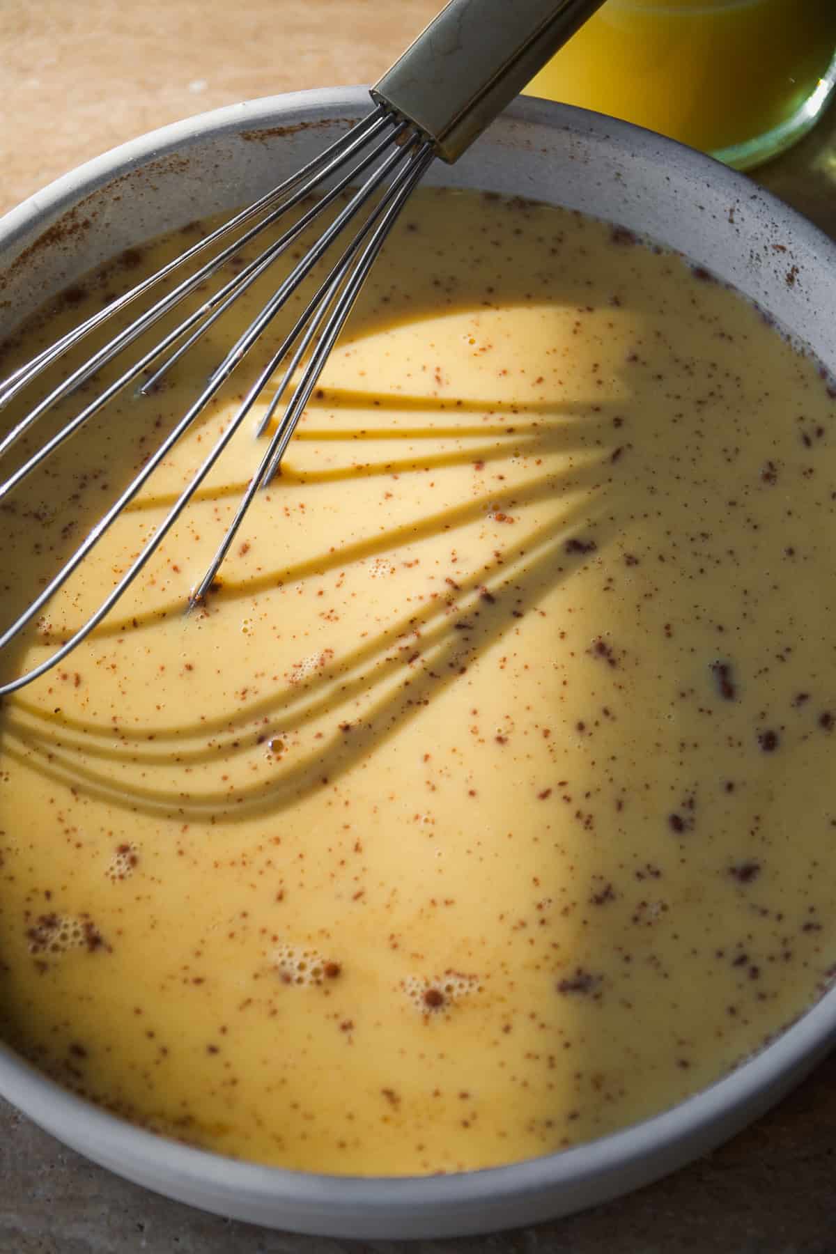 A metal whisk rests in a bowl of creamy yellow sauce or batter with brown specks, illuminated by sunlight. Shadows of the whisk are cast across the surface.