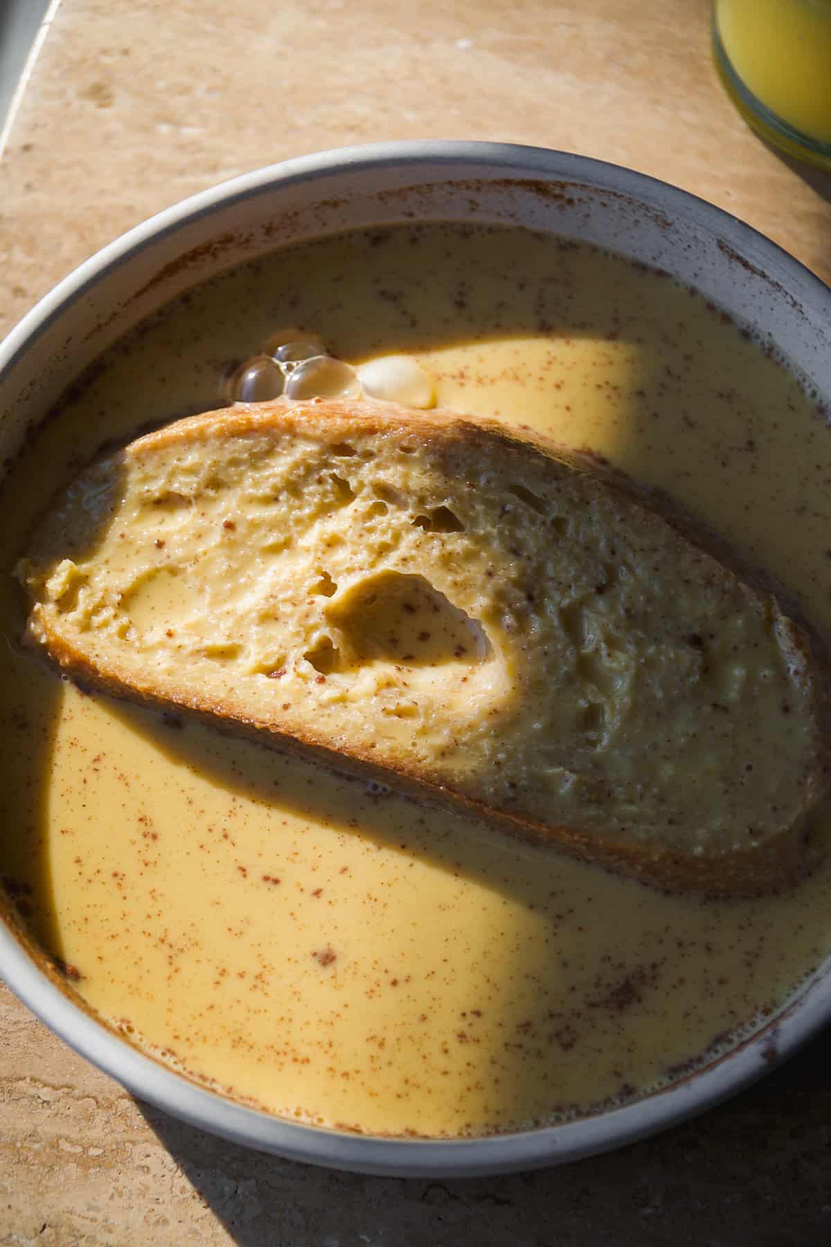 A slice of bread soaks in a creamy, yellow custard mixture with specks of spice, served in a shallow bowl on a light brown surface with some sunlight casting shadows.