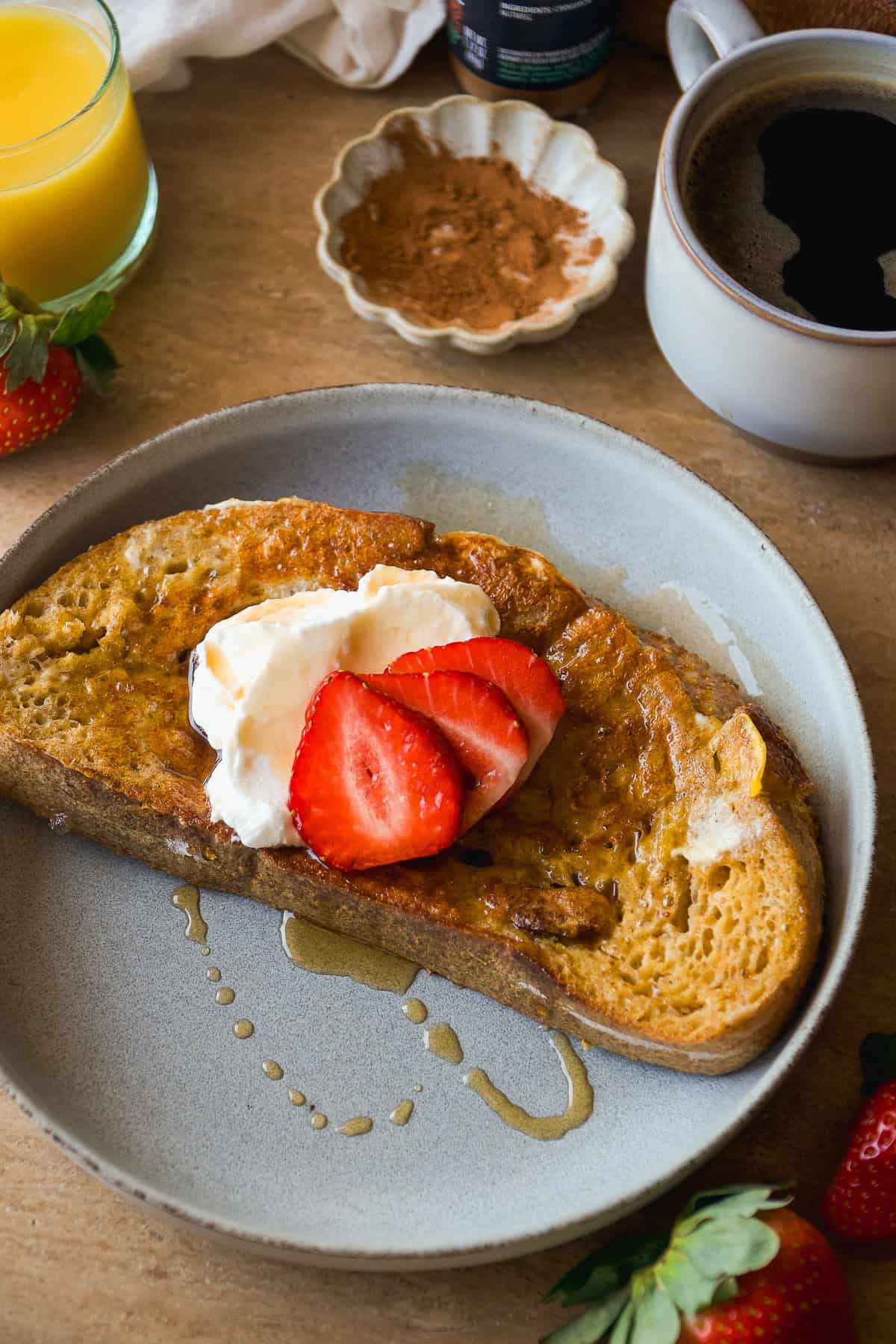 A plate with a slice of French toast topped with whipped cream and sliced strawberries, drizzled with syrup. Nearby are a cup of black coffee, a glass of orange juice, fresh strawberries, and a dish of cinnamon.