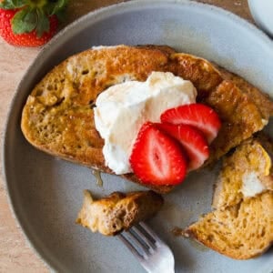 A plate with a slice of French toast topped with whipped cream and sliced strawberries. A fork rests on the plate with a bite of toast. A glass of orange juice, a strawberry, and a bowl of cinnamon are nearby.