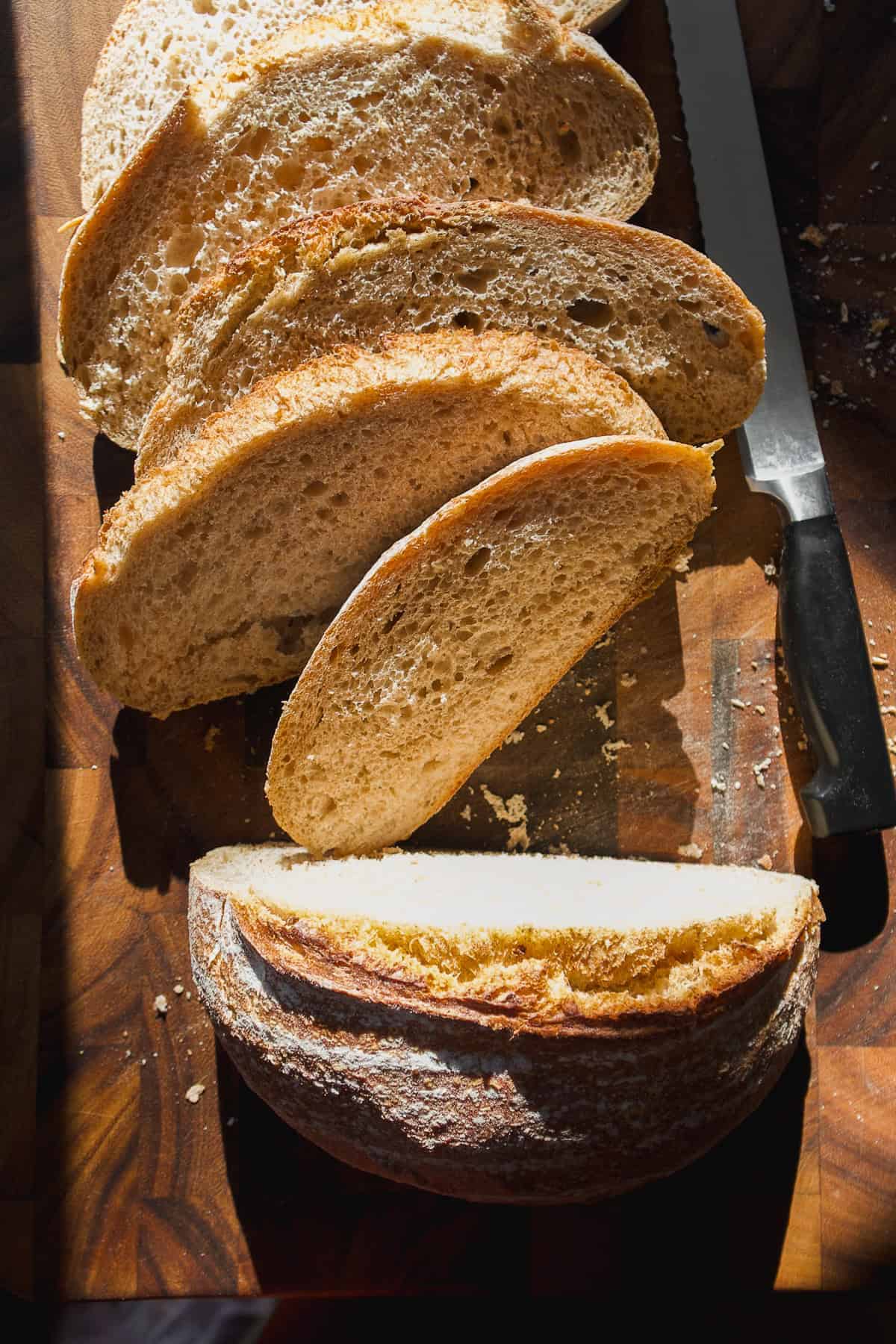 Sliced loaf of rustic bread on a wooden cutting board, with a serrated knife nearby. Sunlight highlights the bread’s golden crust and airy texture.