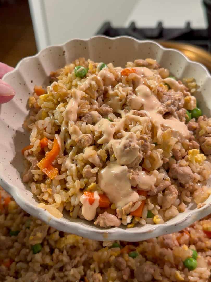 A hand holds a bowl of fried rice with vegetables, ground meat, and a drizzle of creamy sauce on top. The background shows more fried rice in a large serving dish.