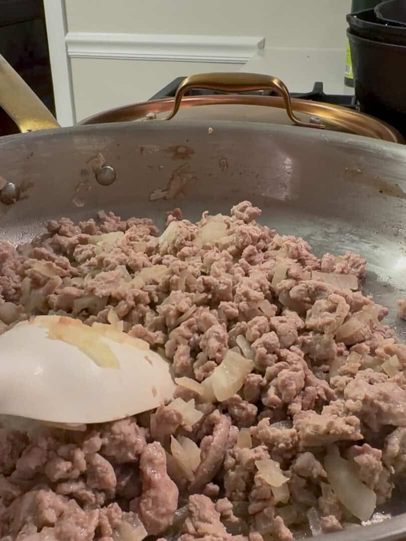 Ground meat and chopped onions are being sautéed in a large, shallow pan, with a white spatula stirring the mixture. The background shows kitchen items, including a bottle and cookware.