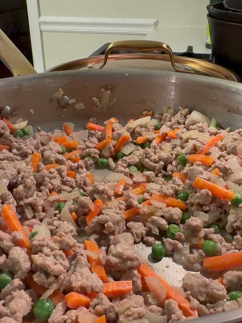 Ground meat is being cooked in a large skillet with diced onions, sliced carrots, and green peas. The mixture is being sautéed on a stovetop, and kitchen utensils are visible in the background.