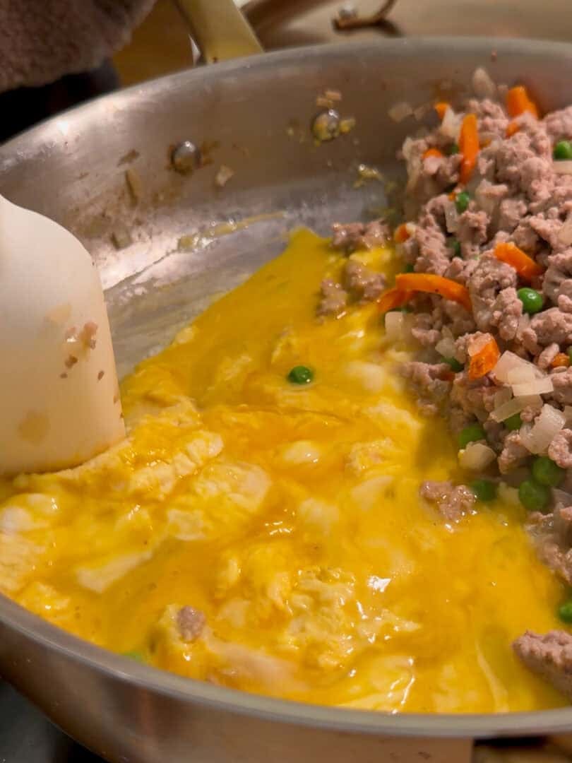 A close-up of a frying pan with scrambled eggs being cooked next to ground meat, carrots, peas, and onions, while a spatula stirs the mixture on a stovetop.