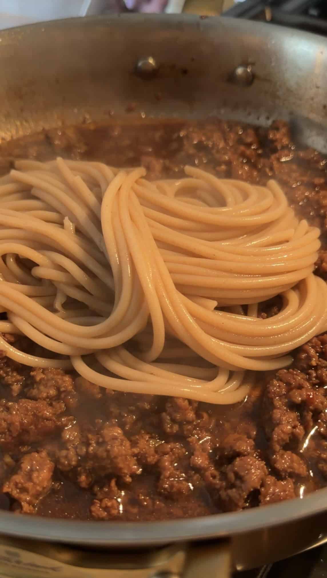 Cooked spaghetti noodles are placed on top of a rich, chunky meat sauce in a pan, with steam rising from the hot mixture.