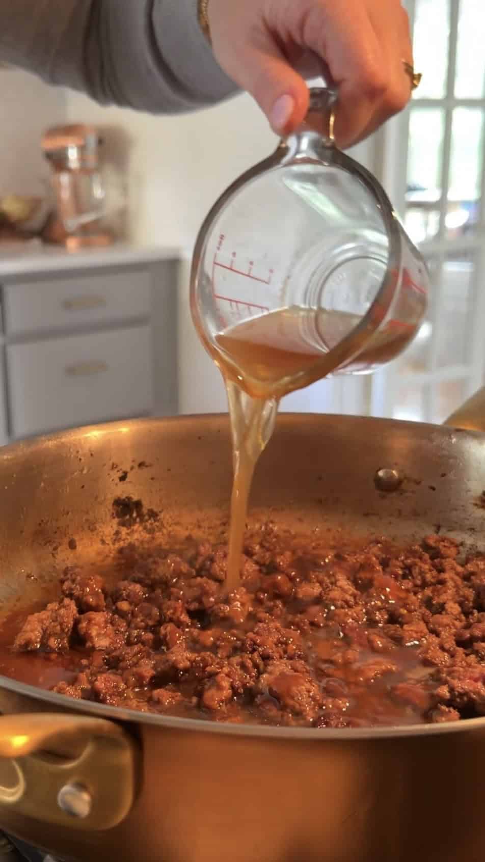 A hand pours broth from a glass measuring cup into a skillet filled with browned ground beef on a stovetop in a kitchen.