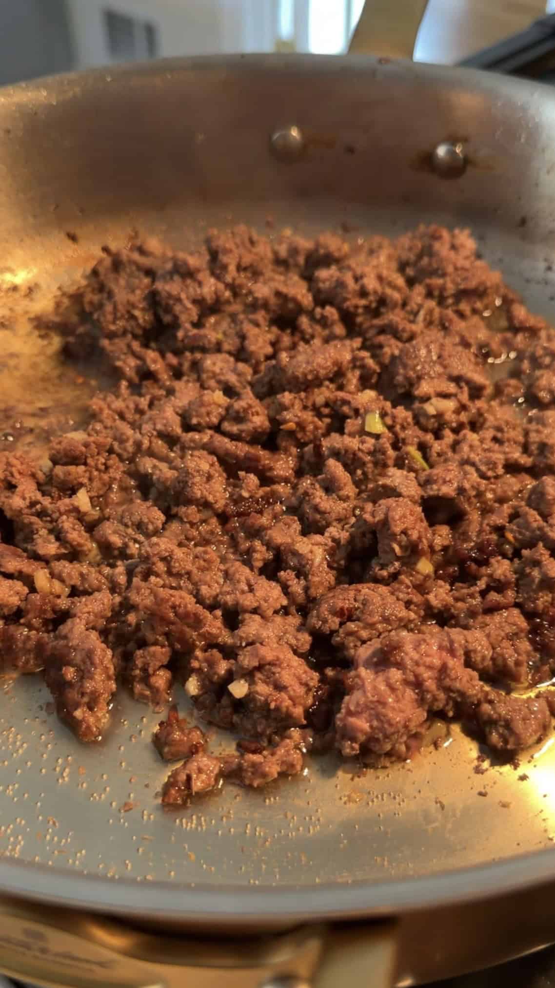 Cooked ground beef browning in a skillet, with small bits of onion visible, glistening with oil under warm light.