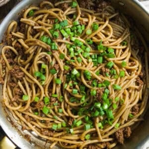 A skillet filled with cooked spaghetti mixed with ground beef and topped with chopped green onions, sitting on a wooden table.
