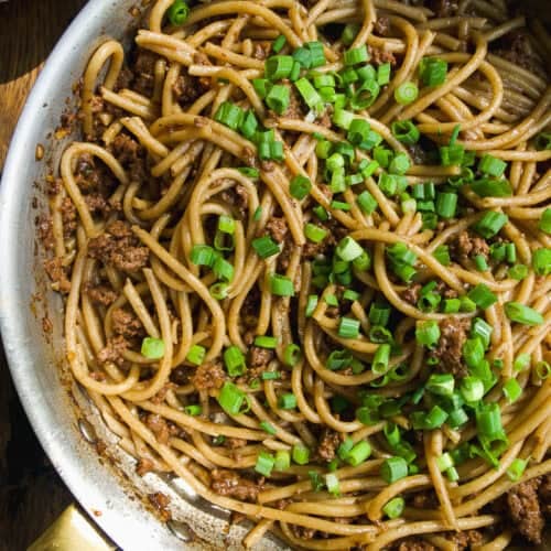 A skillet filled with cooked noodles mixed with ground meat and topped with chopped green onions, sitting on a rustic wooden table.