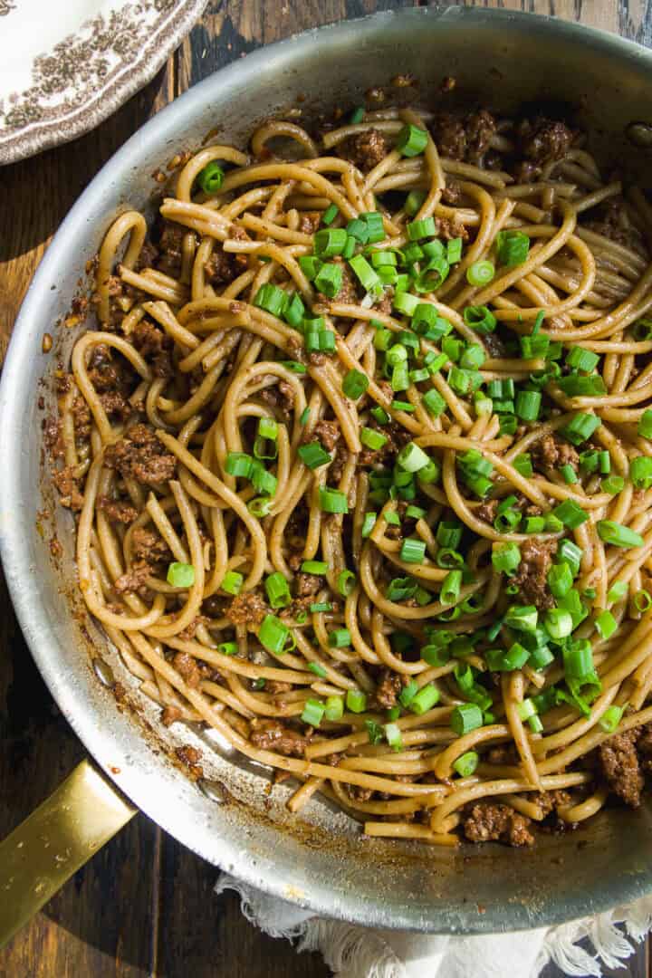 A skillet filled with cooked noodles mixed with ground meat and topped with chopped green onions, sitting on a rustic wooden table.
