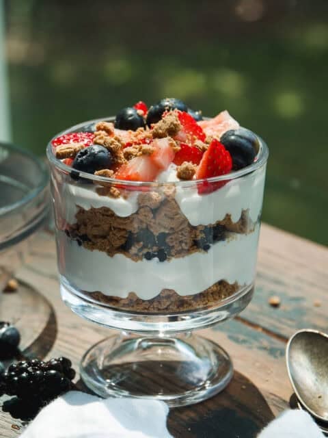 A glass dessert cup filled with layers of whipped cream, crumbled cookies, blueberries, and sliced strawberries, sitting on a wooden table with a spoon and scattered berries nearby.